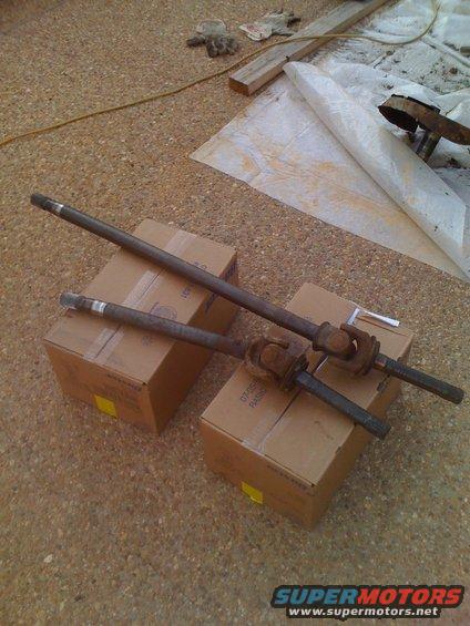 Two drive shafts resting on cardboard boxes on a gravel surface. Some gloves and wood are in the background