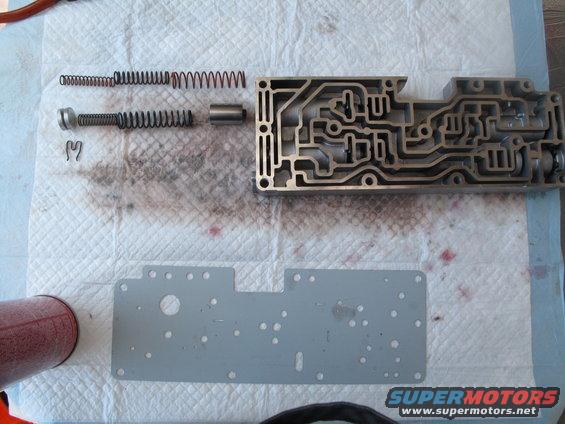 Transmission valve body disassembled with springs and a metal plate on a white absorbent mat. Red spray can visible on left