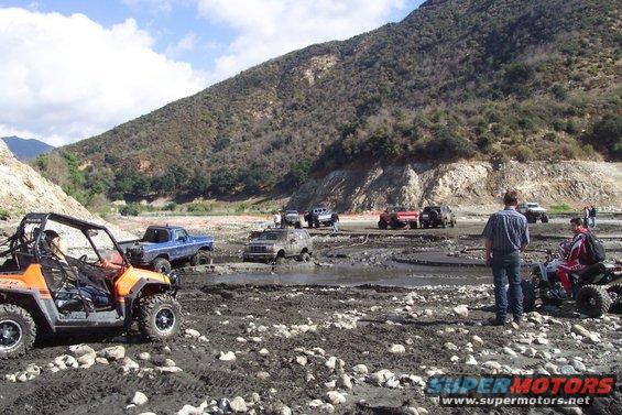 Several off-road vehicles, including a blue pickup truck and a UTV, are in a muddy, rocky riverbed with a large hill in the background
