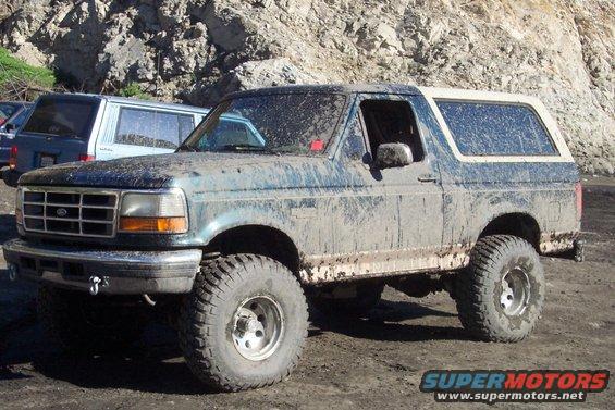 1995 Ford Bronco covered in mud, parked on dirt with rocky hill in background. Another vehicle is partially visible to the left