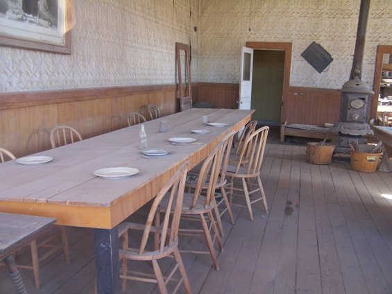 Long wooden table with chairs, set for a meal. Wood stove in corner. Old building interior