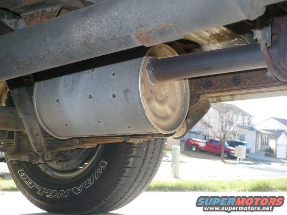 Underside view of a vehicle's exhaust system and tire, with a red pickup truck visible in the background