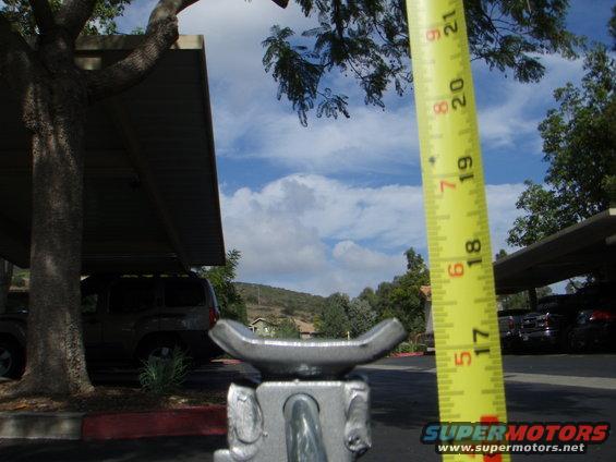 Car jack in foreground with yellow measuring tape, parked SUVs in background under carport