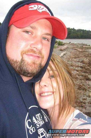 Couple posing for a selfie on a beach with driftwood and water in the background. Man wears a red cap and blue hoodie; woman has blonde hair