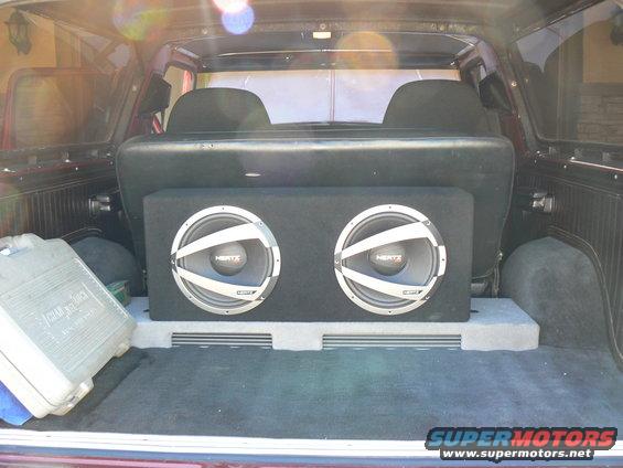 Interior cargo area of a vehicle showing a dual subwoofer box and a grey storage container