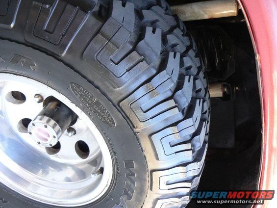 Close-up of a large, aggressive off-road tire mounted on a chrome wheel. Some suspension components are visible