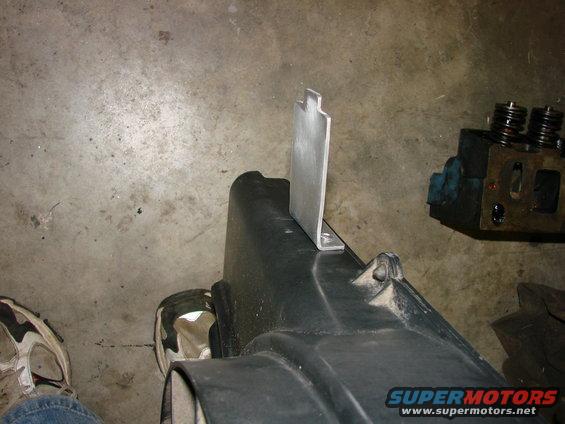 Black plastic engine component with a silver metal bracket attached. Engine block visible in background
