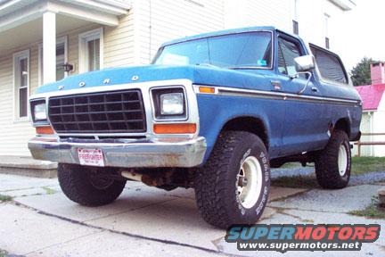 Blue 1978 Ford Bronco with white wheels and off-road tires parked in front of a house