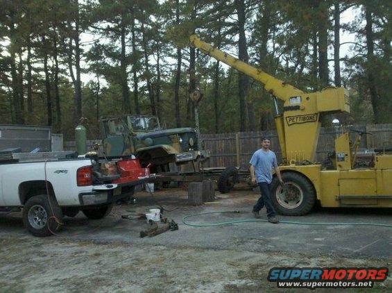 Green military truck on a trailer, being lifted by a yellow Pettibone crane. A white pickup truck is parked nearby