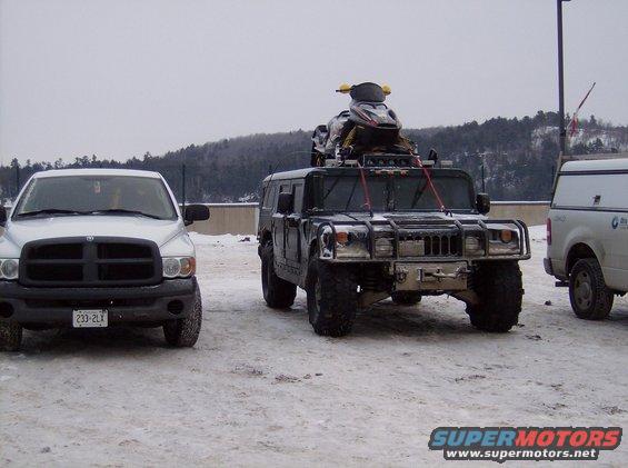 Black Humvee with a snowmobile on its roof rack, parked in snow next to a white pickup truck