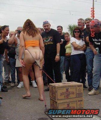 Woman in orange top and leopard print thong, barefoot, is led by a leash attached to stacked beer boxes