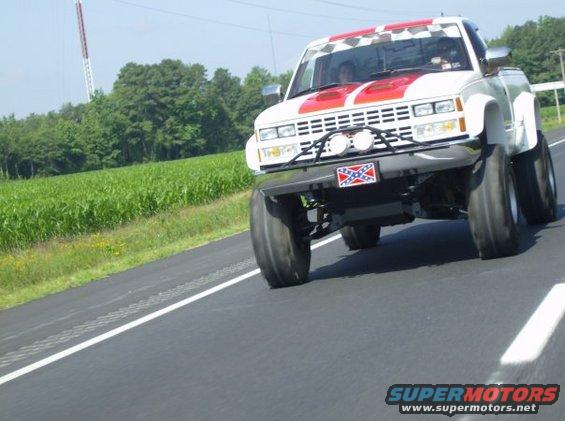 Lifted white pickup truck with red racing stripes and checkerboard pattern on the windshield banner, driving on a road next to a cornfield