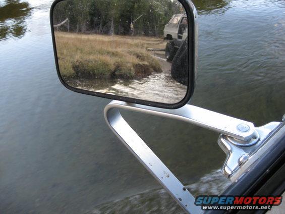 Side mirror reflection of a light-colored truck with large tires driving on a grassy bank near water