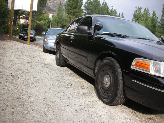 Black 2004 Ford Crown Victoria police car in foreground, light blue sedan behind it on gravel shoulder