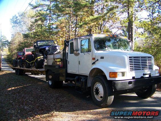 White flatbed truck with a trailer carrying two jeeps. The truck has '2000' on the fender