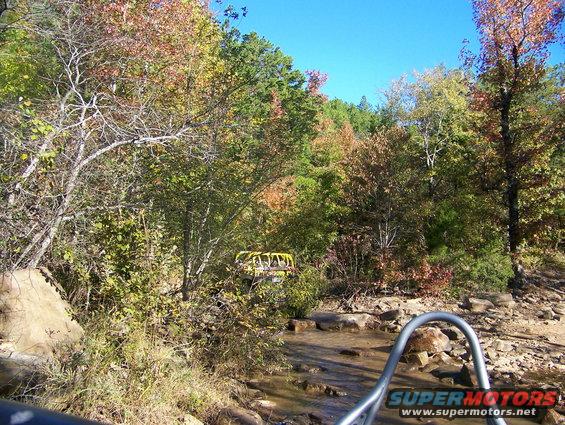 Yellow off-road vehicle partially visible among trees and rocks near a stream