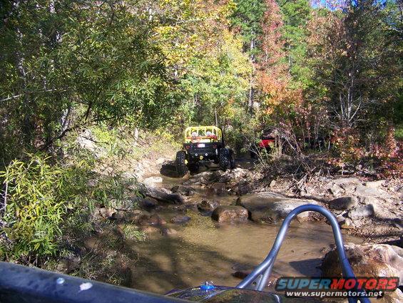 Two off-road vehicles navigating a shallow, rocky stream in a wooded area with fall foliage