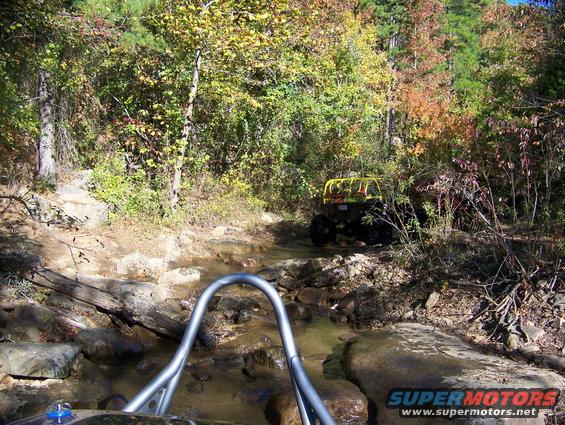 Off-road vehicle with yellow roll cage driving through a rocky stream