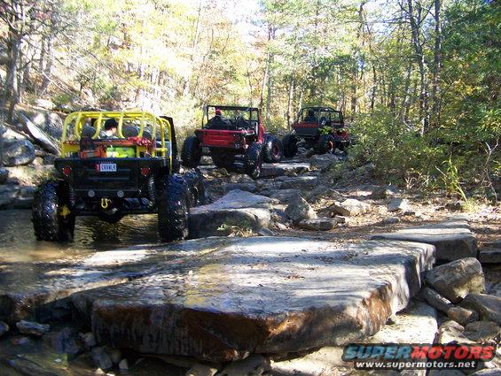 Three off-road vehicles navigate a rocky stream bed with yellow roll cages