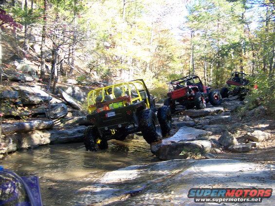 Several off-road vehicles, including a yellow-caged Jeep, are driving over rocks in a stream. Another red Jeep is behind it, and a third vehicle is further back