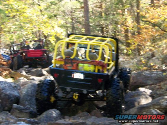 Rear view of black off-road vehicle with yellow roll cage on rocky trail; another red vehicle behind