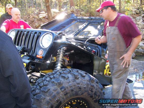 Jeep Wrangler front end with hood open, people gathered around. Large off-road tires visible