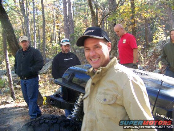 Close-up of a modified off-road vehicle's suspension and tire, with people in the background