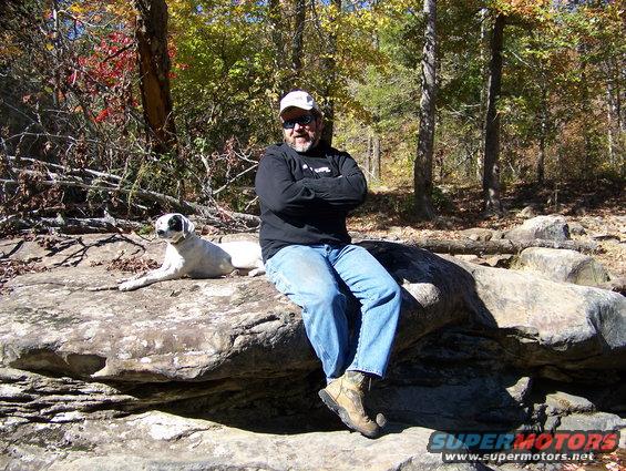 man with arms crossed and dog sitting on large rocks in woods, fall foliage visible