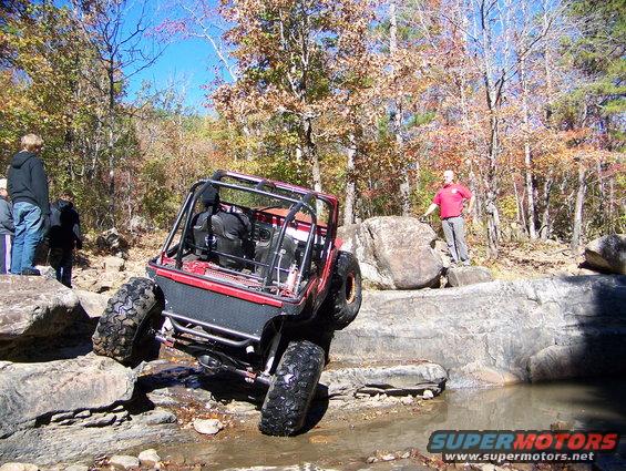 Red off-road vehicle with roll cage climbing over rocks near water
