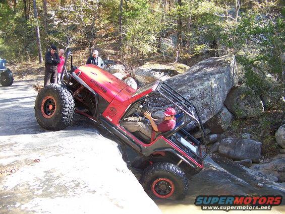 Red Jeep Wrangler off-roading over rocks, driver steering through water