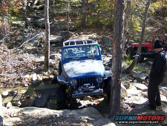 Blue Jeep Wrangler going over rocks, red Jeep in background, person pointing