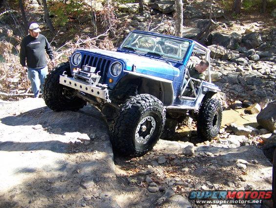 Blue Jeep Wrangler with large tires and winch on rocky terrain, driver inside