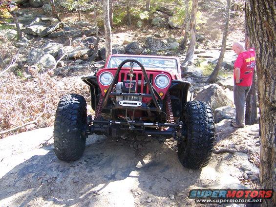 Red Jeep Wrangler with large tires on a rocky trail, man standing nearby