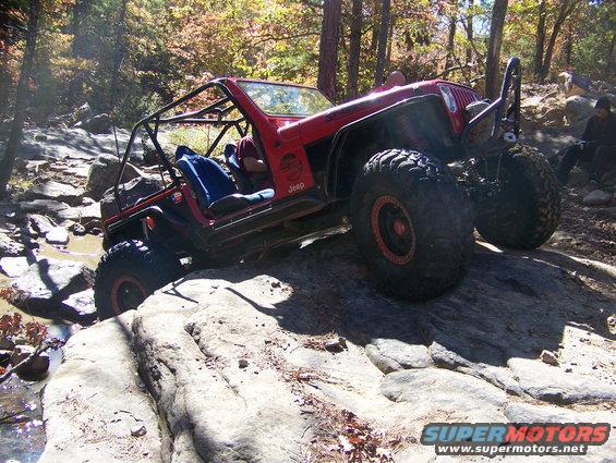 Red Jeep Wrangler with roll cage and large tires navigating rocky terrain