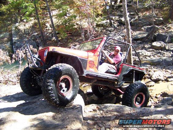 Red Jeep Wrangler with large tires navigating rocky terrain. Driver wearing a red cap and shirt