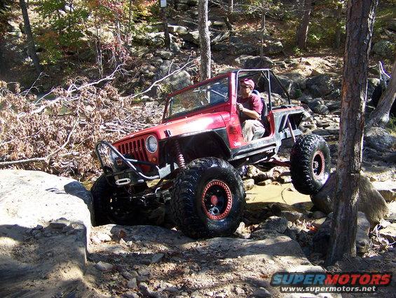 Red Jeep Wrangler with large tires navigating rocky terrain, driver visible