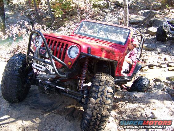 Red Jeep Wrangler with custom roll cage and winch, navigating rocky terrain. Another off-road vehicle is visible in the background