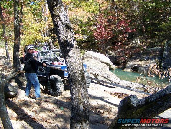 Person pulling a strap attached to a blue off-road vehicle near a creek. Another off-road vehicle is behind it