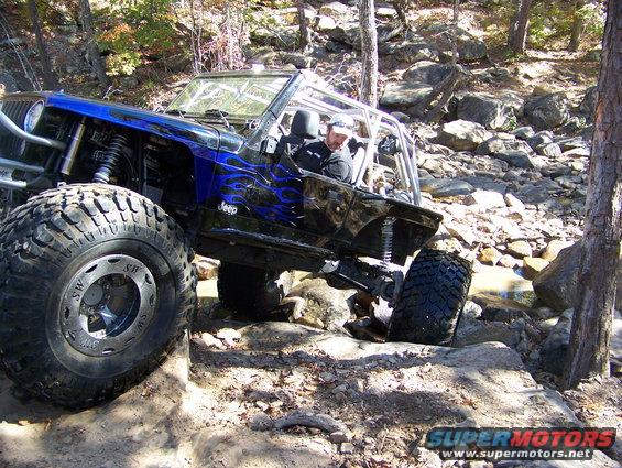 Modified Jeep Wrangler with large tires navigating rocky terrain. Blue flame decals on the side