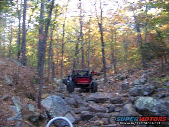 Red off-road vehicle navigating a rocky trail in a forest with autumn foliage