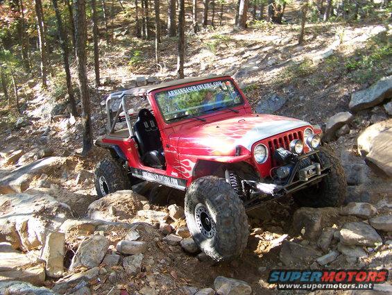 Red Jeep Wrangler with flame decals and off-road tires navigating a rocky trail. Text on windshield reads 'ARKANSASSCRAWLERS.COM'