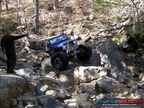 Blue off-road vehicle with large tires navigates a rocky trail, a person points ahead