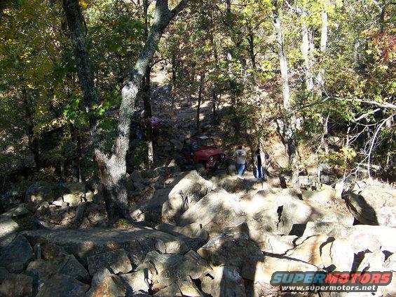 Red off-road vehicle on rocky trail with trees and people