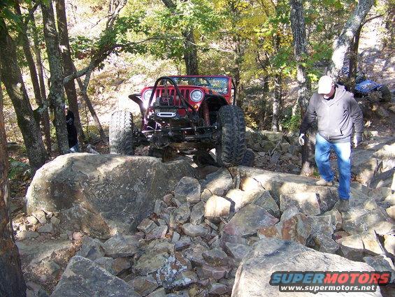 red jeep on rocks, guy in white hat walking nearby, another vehicle in background