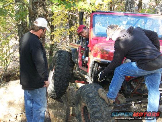 Red Jeep Wrangler with large tires on rocky terrain, people working on engine