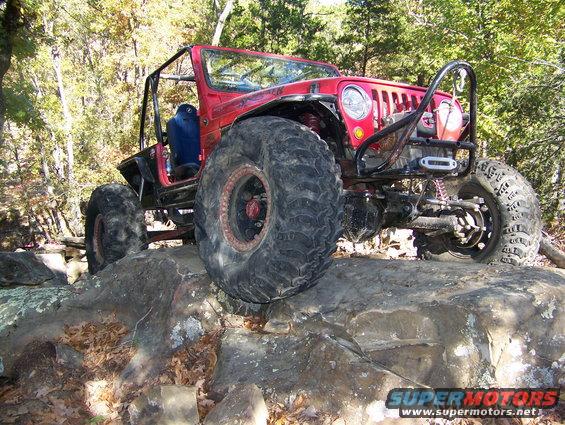 Modified red Jeep with large tires climbing over rocks. Winch visible on front bumper