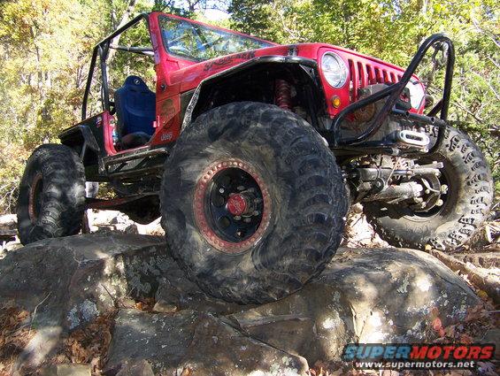 Red Jeep Wrangler with large tires on rocks. Has a roll cage and winch