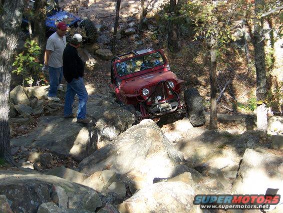 Red Jeep Wrangler navigating rocky terrain with two men watching