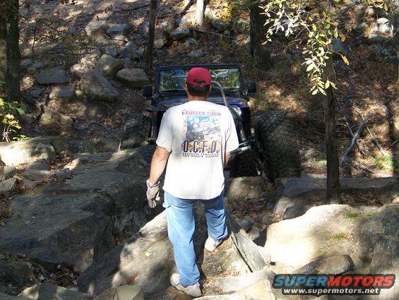 guy in red hat and white shirt watching a blue off-road vehicle crawl over rocks