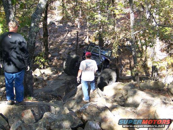 Off-road vehicle navigating a rocky trail, with two men observing